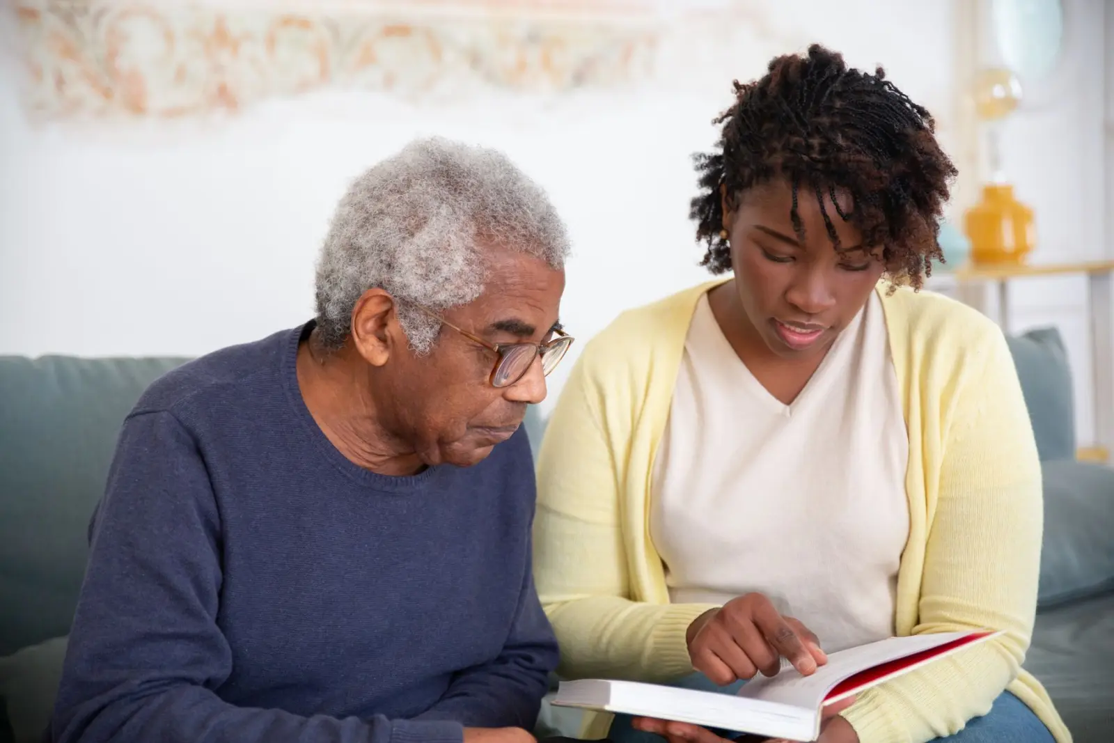 A caregiver and an older man reading a book together on a living room sofa